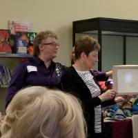 A woman holds a shadow box while a second woman points to a spot behind it